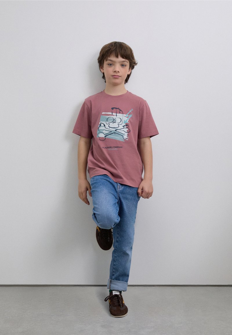 Różowy graficzny t-shirt z nadrukiem kreskówkowej czaszki z skrzyżowanymi kośćmi. Noszony z niebieskimi dżinsami i brązowymi trampkami. Minimalistyczne tło.