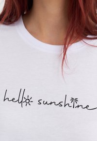 Vit t-shirt med texten "hello sunshine" i svart, kursiv stil tillsammans med en liten sol- och palmträdsgrafik.