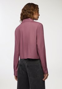 Camicia a maniche lunghe, cropped, in morbido tessuto malva, con colletto classico e polsini con bottoni, abbinata a jeans grigio scuro a vita alta.