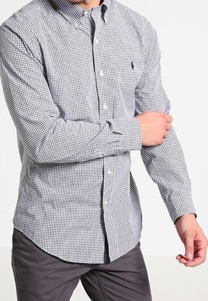 Mand, der justerer manchet på langærmet sort-og-hvidternede skjorte, iført mørkegrå bukser, foran hvid baggrund.