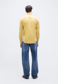 Άνδρας που στέκεται με την πλάτη προς τα εμπρός φορώντας κίτρινο πουκάμισο με μακριά μανίκια, μπλε τζιν και σκούρα παπούτσια σε απλό λευκό φόντο.