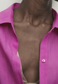 Camicia di lino fucsia con una consistenza morbida, caratterizzata da un colletto aperto e un singolo bottone rosa. Indossata sopra un top beige.