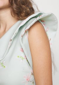 Vaaleanvihreä kukkakuosiset mekko, jossa on frillasivat olkapäädetailit ja sileä pinta. Lähikuva olkapäästä ja käsivarresta.