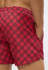 Red swim shorts featuring a repeating "HUGO" logo pattern in black, with an elastic waistband and side pockets. Textured fabric.