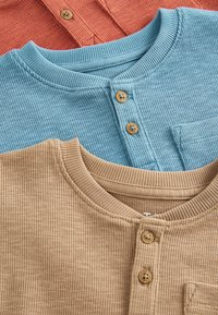 Trzy prążkowane koszulki henley w jasnoniebieskim, koralowym i beżowym kolorze. Każda z nich ma okrągły dekolt, drewniane guziki oraz kieszeń z przodu beżowej koszulki.