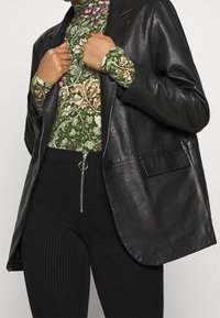 Svart skinnkavaj med slag, sidofickor och en knapp. Grön mönstrad polotröja med blommönster, kombinerad med svart randiga byxor.