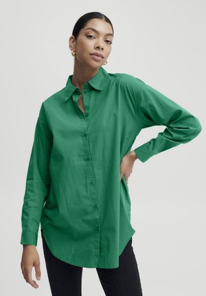 Grüne, knöpfbare Bluse aus weicher Baumwolle, mit einem spitzen Kragen, langen Ärmeln und einem leicht geschwungenen Saum. Getragen zu dunkelblauen Jeans.