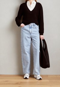 Schwarzer V-Ausschnitt-Pullover, weißes Hemd mit Kragen, hellblaue gerade Jeans, weiße Sneakers und eine schwarze Tragetasche. Glatte Texturen, lässige Passform.