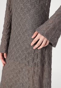 Vestido cinza de renda com mangas longas, apresentando padrões florais e uma textura transparente. Close-up destaca os detalhes intrincados e a colocação das mãos.