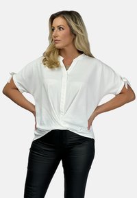 Weiße kurzärmelige Bluse mit Knopfleiste und geknotetem Vorderdetail. Hergestellt aus weichem Stoff mit lockerer Passform. Getragen mit schwarzen Hosen.