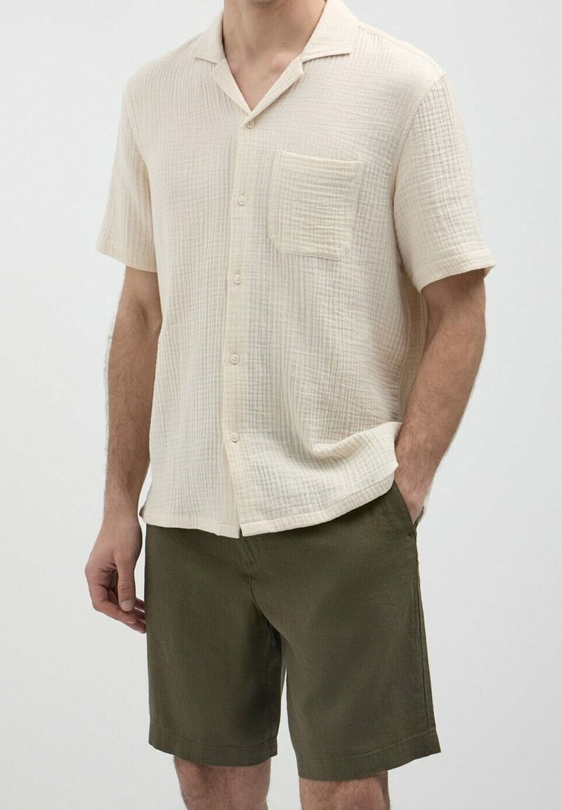 Homme portant une chemise crème à manches courtes avec une poche poitrine et un short vert olive, debout avec une main dans la poche.