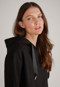 Felpa nera con cappuccio e coulisse con punte metalliche, tessuto texturizzato e design classico. Il modello ha i capelli lunghi e mossi e uno sfondo neutro.