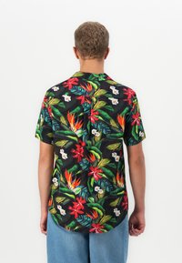 Man in een kortebloes met zwarte kleur en kleurrijke tropische bloemenprint, gecombineerd met lichtblauwe jeans, gezien van achteren.