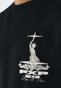 Zwarte stof met zilveren grafiek van een gestileerd standbeeld dat sterren vasthoudt, met de teksten "PXP," "Change the Game," en "Project X Paris."