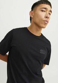 Schwarzes Baumwoll-T-Shirt mit kurzen Ärmeln, Rundhalsausschnitt, mit einem strukturierten Logo in Dunkelgrau auf der linken Brustseite. Glatte Stoffqualität.