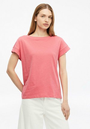 Młoda kobieta z długimi włosami, ubrana w różowy t-shirt z krótkim rękawem i białe spodnie na jednolitym białym tle.