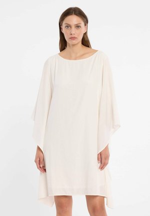 Woman wearing a loose, knee-length white dress with wide sleeves and a round neckline, standing against a plain white background.