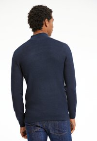 Tumma sininen neulepaita pitkillä hihoilla, korkealla kauluksella ja ribatulla helmalla. Sujuva rakenne, jossa on hienoinen venyvyys, näkyy takaa.