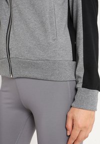 Sweat-shirt zippé gris avec des panneaux latéraux noirs, des poignets et un ourlet côtelés. Fabriqué dans un tissu doux avec une texture lisse, idéal pour les vêtements de sport.