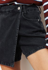 Shorts en denim noir avec une coupe décontractée, poches avant et un design unique avec fente sur le côté. Tissu texturé avec un léger éclat.