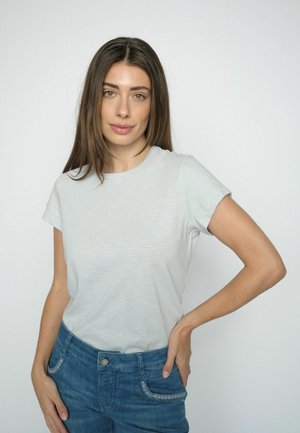 Fiatal nő hosszú barna hajjal, világos csíkos pólót és kék farmert visel, egyik kezét a csípőjén tartva áll egyszínű háttér előtt.