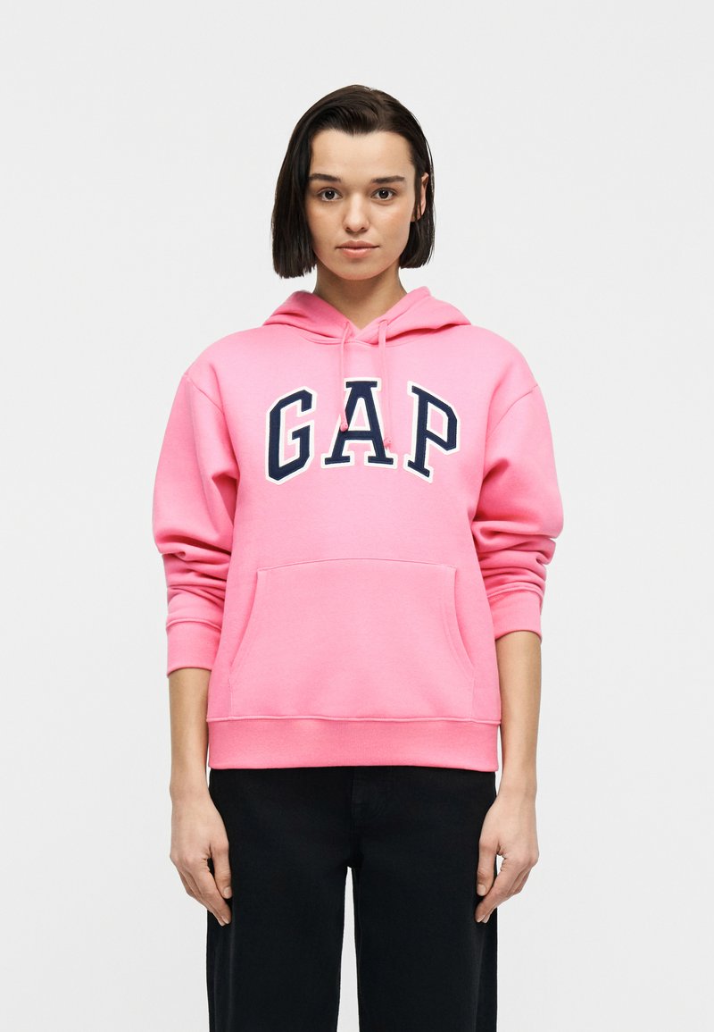 Νεαρό άτομο με κοντά σκούρα μαλλιά, φοράει ένα φωτεινό ροζ φούτερ με μεγάλο λογότυπο «GAP» και μαύρο παντελόνι, στέκεται μπροστά σε απλό φόντο.