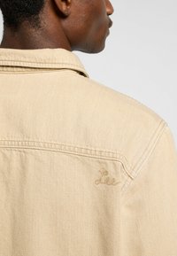 Chaqueta de mezclilla beige con cuello puntiagudo, costura en el yugo trasero y un sutil logo bordado en el hombro izquierdo.