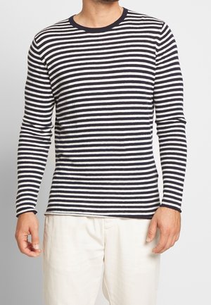 Mand iført en langærmet marineblå og hvid T-shirt med horisontale striber og off-white bukser, stående foran en ensfarvet baggrund.