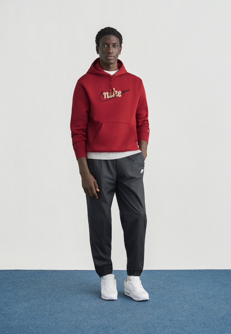 Felpa con cappuccio rossa con logo Nike dorato, maglietta grigia sotto, abbinata a pantaloni neri affusolati e sneakers bianche. Design semplice con polsini elastici.