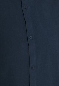 Chemise boutonnée bleu marine en tissu lisse et léger. Présente une patte droite avec quatre boutons sur le devant.