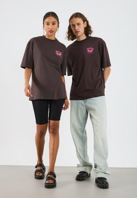Brown oversized t-shirts feature pink graphic print. Model wears black bike shorts and black sandals; second model in light blue jeans and black shoes.