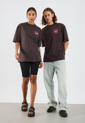 Braune übergroße T-Shirts mit pinkem Grafikdruck. Das Model trägt schwarze Radlerhosen und schwarze Sandalen; das zweite Model ist in hellblauen Jeans und schwarzen Schuhen.