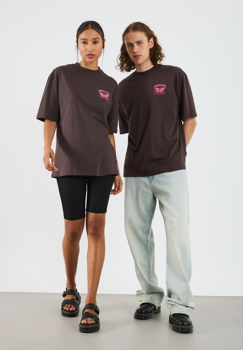 Brown oversized t-shirts feature pink graphic print. Model wears black bike shorts and black sandals; second model in light blue jeans and black shoes.