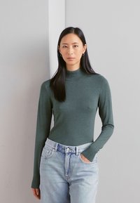 Dolgimi rokavi, oprijet turtleneck v temno zeleni pletenini, v kombinaciji z modrimi kavbojkami z visokim pasom. Preprost dizajn, minimalne detajle.
