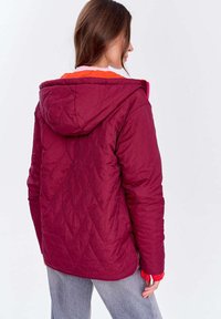 Kobieta ubrana w kapturową, pikowaną, ciemnoczerwoną kurtkę i szare dżinsy, pokazana od tyłu na jednolitym jasnym tle.