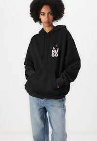 Svart hettegenser med frontlomme, snorhette og brodert "HUGO"-logo i forskjellige farger på brystet.