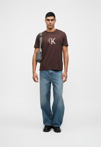 Hnedé bavlnené tričko s logom, voľný strih svetlomodré džínsy a čierne topánky. Sivá crossbody taška so popruhom cez rameno.