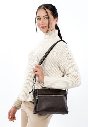 Bruine leren crossbodytas met een gestructureerde afwerking, ritssluiting en verstelbare band, gedragen door een persoon die een crèmekleurige trui draagt.