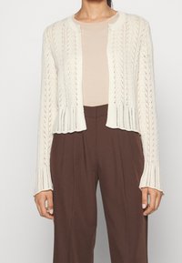 Pletený kardigan v off-white barvě, s otevřeným předním dílem, texturovaným vzorem a dlouhými rukávy. V kombinaci s béžovým topem a hnědými kalhotami.