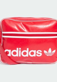 Borsa sintetica rossa con dettagli bianchi, dotata di tasca frontale con zip, grande logo "adidas" e iconico logo trefoil. Tracolla regolabile.