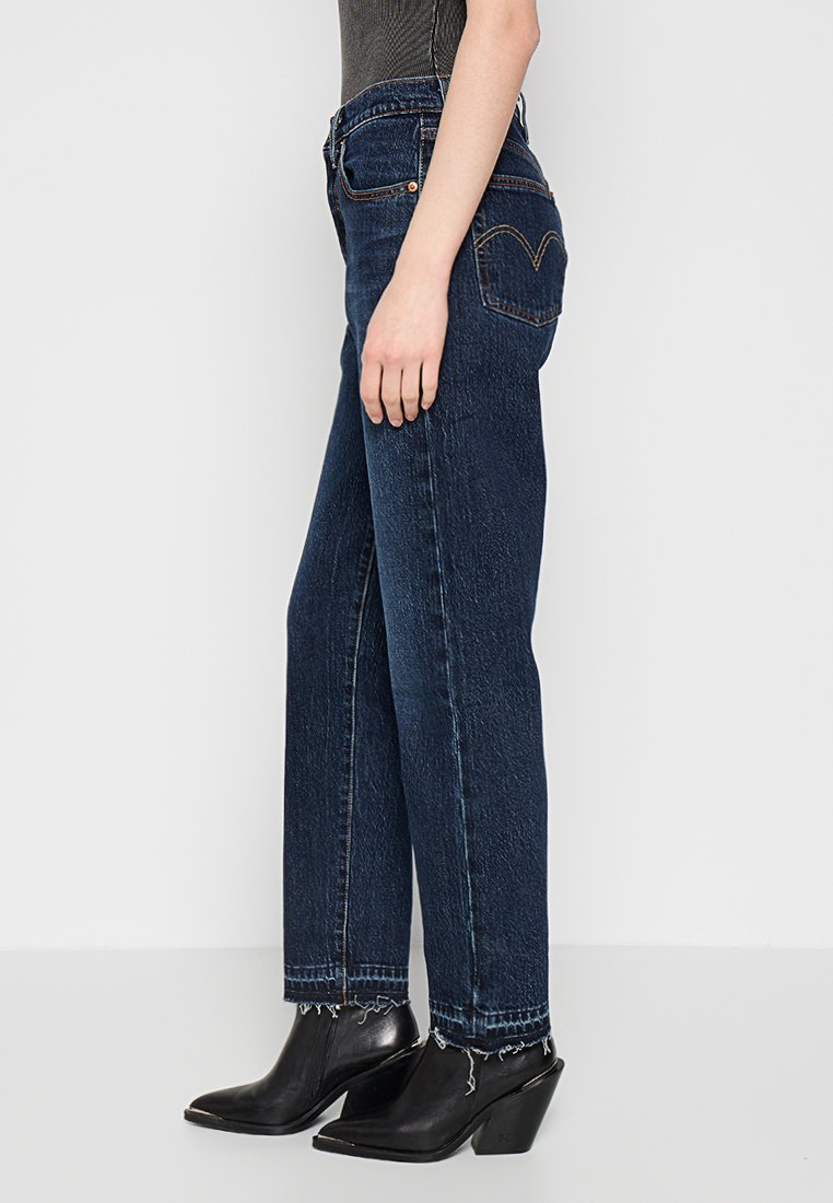 Jean taille haute bleu foncé avec un design à jambe droite, présentant un léger délavage et un motif de couture distinctif sur les poches arrière. Portés avec des bottes à talons noires.