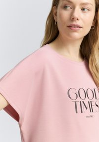 Rožinė per didelė palaidinė su apvaliu iškirpimu, trumpomis rankovėmis ir tamsiai pilku grafiniu spaudiniu, kuriame užrašyta "GERI LAIKAI nuo 1962".