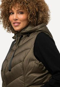 Olivově zelená prošívaná vesta s kapucí a stahovací šňůrkou, nošená přes černé tričko s dlouhým rukávem, s předním zipem a bočními švy.