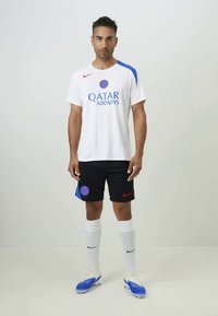 Man standing in white soccer jersey and socks, black shorts, and blue cleats against a plain light background.