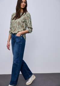 Femme aux longs cheveux foncés portant un chemisier vert à motifs, un jean bleu à jambes larges et des baskets blanches, debout avec une main dans la poche.