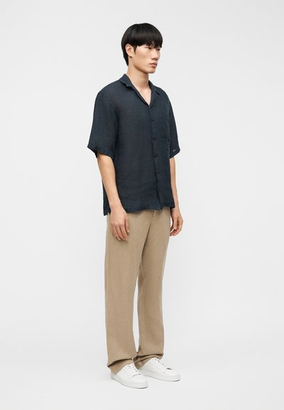 Σκούρο μπλε πουκάμισο από λινό με κοντά μανίκια και τσέπη στο στήθος, συνδυασμένο με ανοιχτό μπεζ παντελόνι από λινό και λευκά αθλητικά παπούτσια, στέκεται μπροστά σε ένα απλό φόντο.