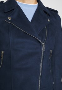 Mörkblå jacka i konstmocka med dubbelknäppt design, silvriga dragkedjor och tryckknappsdetaljer. Texturerat tyg med detaljerad sömnad.