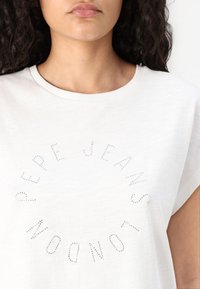 White cotton t-shirt with a round neckline. Features embossed rhinestones forming the text "PEPE JEANS LONDON" in a circular pattern.