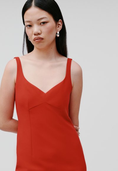 Rode mouwloze jurk met een diepe V-hals en een getailleerde silhouet. Glad materiaal, minimale naden en een strakke design. Zilveren creolen.