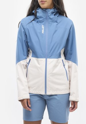 Veste bicolore bleu clair et crème avec un col montant, une fermeture éclair à l'avant et deux poches latérales. Portée avec un short bleu assorti.